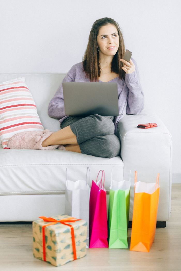 A woman sits comfortably on a sofa, using a laptop for online shopping with shopping bags nearby.
